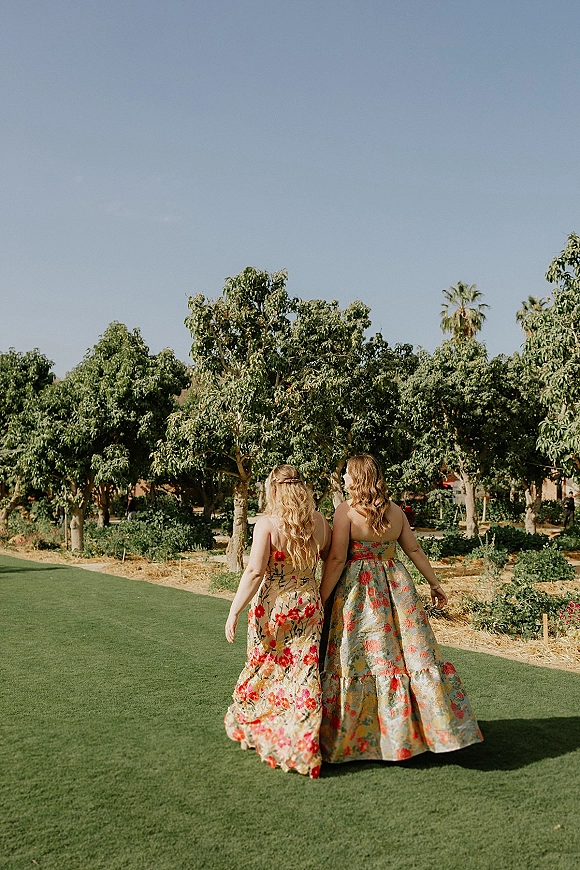 Wedding guest dresses in floral print as two guests walk along a garden path on a grass lawn with orchard trees and clear sky