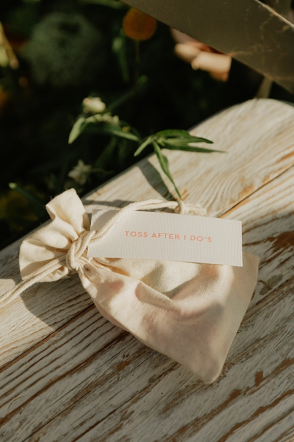 Wedding favor bag with muslin favor bags drawstring pouch, twine-tied tag and greenery sprig on a rustic wooden table with blurred foliage
