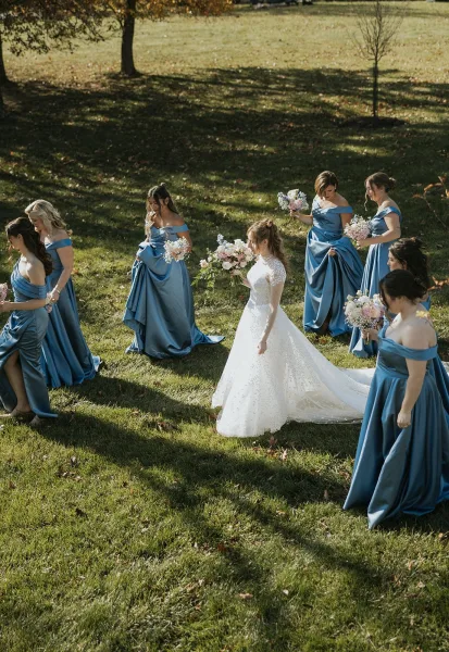 Bride with bridesmaids in blue dresses walking on a sunlit lawn, her lace-sleeved gown and long train trailing behind pastel bouquets with baby's breath