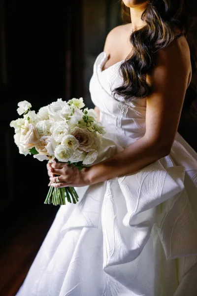 Bridal portrait with bridal bouquet white roses, bride in a strapless gown holding greenery stems by soft window light in a dark interior