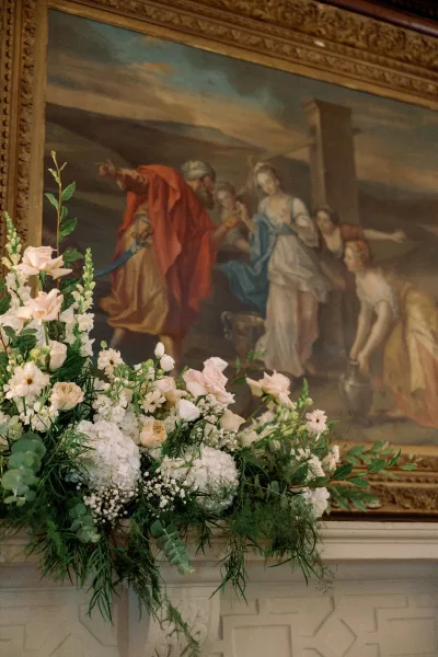 Wedding floral arrangement of white hydrangea and blush roses with greenery on a mantel beneath an ornate gold-framed painting