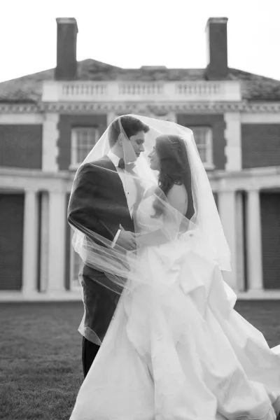 Couple portrait in a black and white wedding portrait, bride and groom embracing under a veil on the lawn before a brick mansion