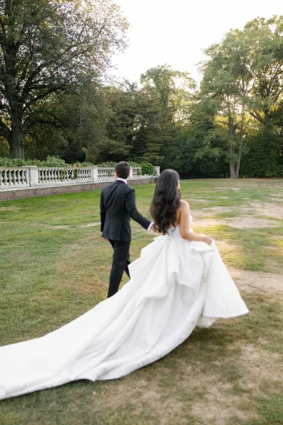 Couple portrait of bride and groom walking away holding hands, her long wedding dress train trailing on a lawn by stone balustrade