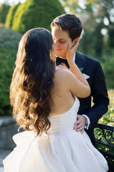 Wedding couple portrait of bride and groom embrace, touching foreheads in a sunlit garden by a wrought iron bench, strapless gown and tuxedo