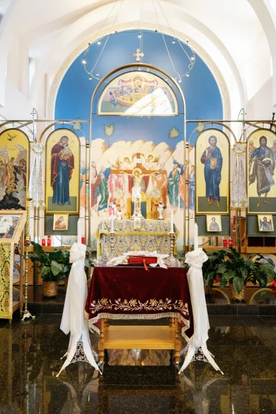 Church ceremony altar with red velvet altar cloth, candles and icons, set before a blue-walled iconostasis under chandelier lights