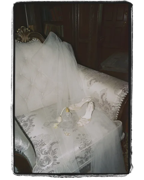 Bridal accessories arranged in a wedding detail flat lay with veil, ivory heels, perfume bottle, and rings on a settee by bookshelves