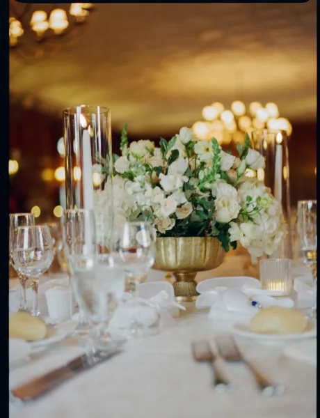 Reception tablescape with wedding table centerpiece of white florals and greenery in a gold compote, surrounded by glowing candles under warm bistro lights