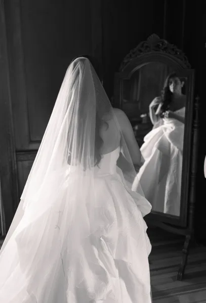 Bridal portrait of a bride looking in mirror, holding her full skirt gown with a veil in a vintage wood-paneled bedroom interior