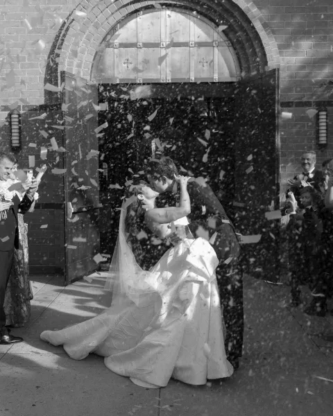 Wedding kiss during confetti exit as groom dips the bride in a veil outside a brick church, guests cheering by arched doors