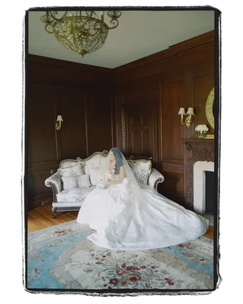Bridal portrait of a bride sitting on sofa in a strapless gown with long train, holding a bouquet in a wood-paneled room by the fireplace
