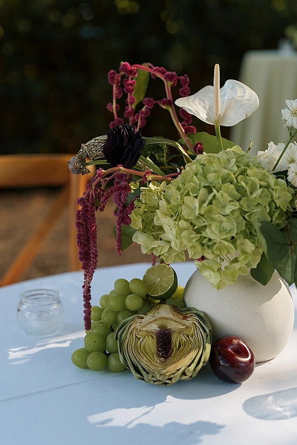 Wedding centerpiece with hydrangea and anthurium in a ceramic vase, with green grapes, lime, plum, and glass votive on white tablecloth outdoors