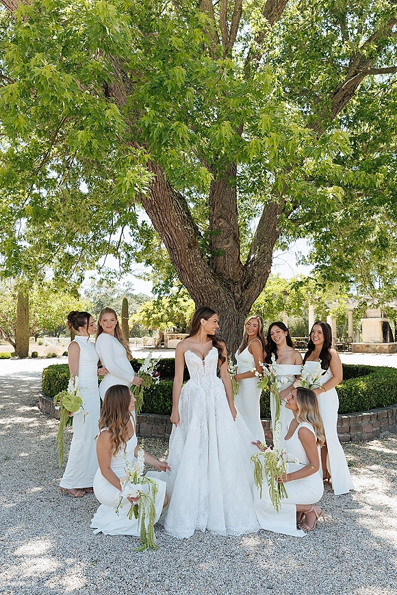 Bridesmaid group photo with bride and bridesmaids in white dresses holding long-stem bouquets on a sunlit garden courtyard path under a large tree