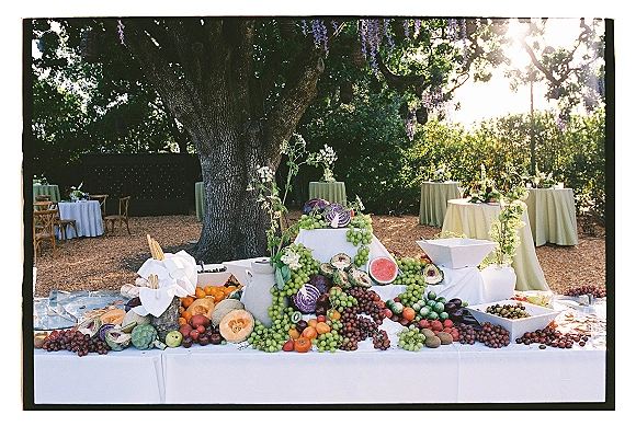 Wedding dessert table with a fruit wedding dessert table display of grapes, watermelon and citrus under a tree canopy in a sunlit garden setting