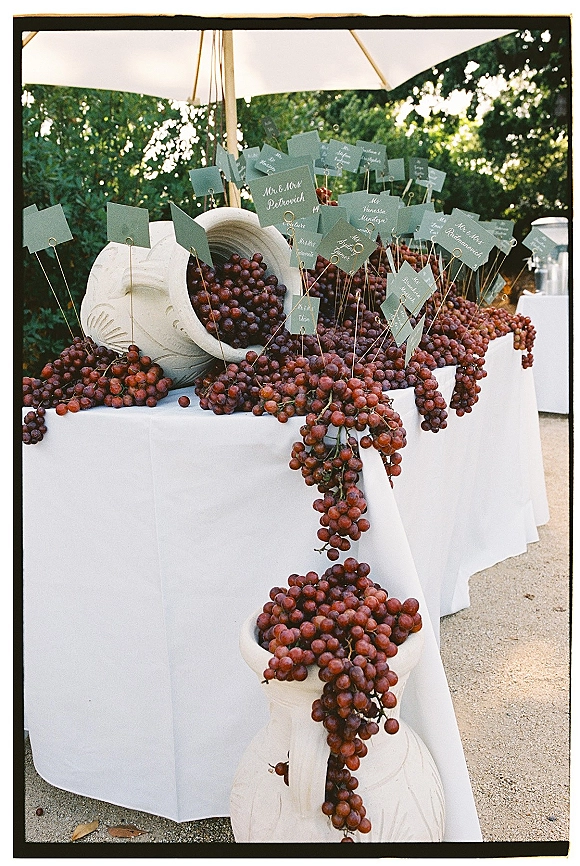 Escort card display with wedding escort cards in calligraphy, hanging card picks and cascading grapes on stone urns over a white linen table outdoors
