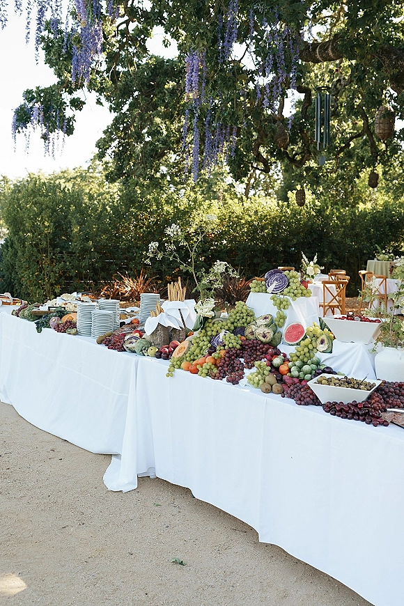 Wedding dessert table with a wedding fruit display of grapes, watermelon, figs, and cheese boards on white linen under a wisteria-draped tree