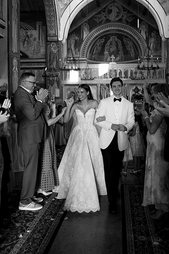 Wedding recessional as bride and groom walk the aisle under chandeliers, confetti petals falling as guests film in an icon-filled church interior