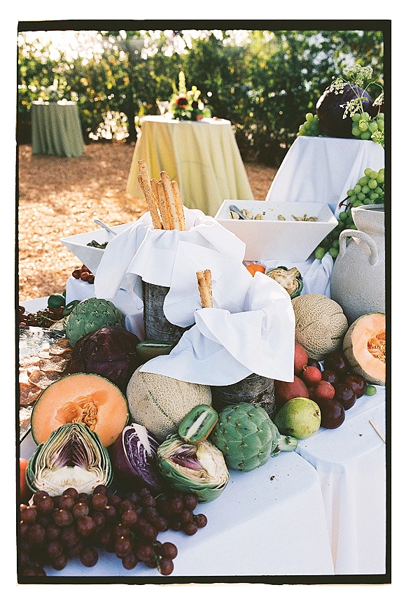 Wedding appetizer table with a wedding cocktail hour spread of breadsticks, crackers, grapes, melon, and citrus on white linens in a garden setting