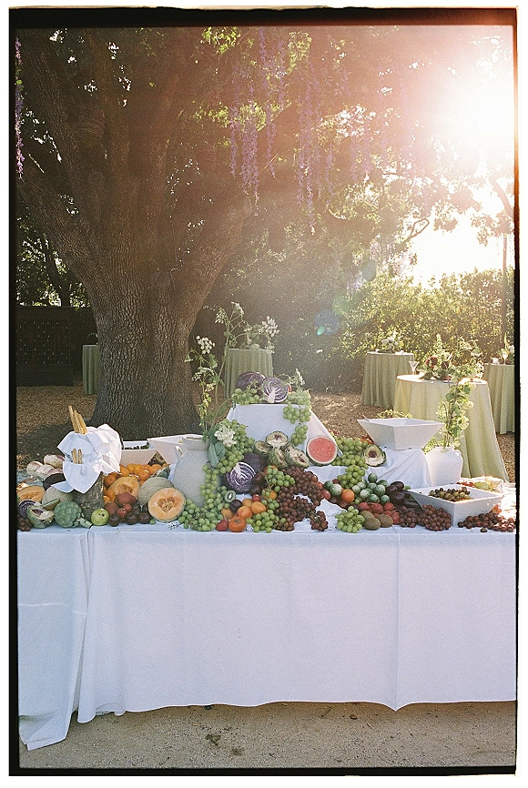 Wedding fruit display with grapes, citrus, watermelon and figs arranged in bowls on a white linen table under sunlit garden trees
