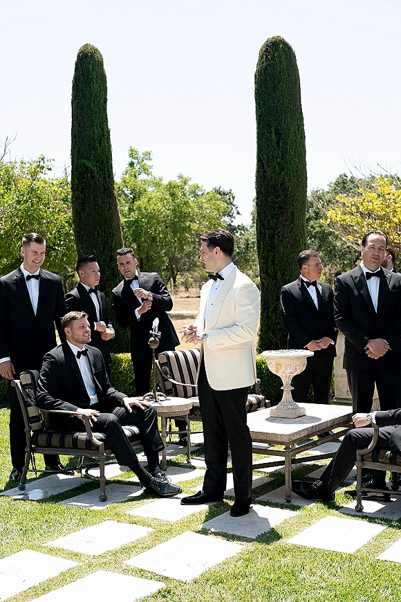 Groomsmen portrait of groom in white tuxedo with groomsmen in black tuxedos lounging by patio chairs on a garden lawn under cypress trees