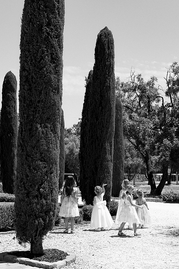 Flower girls in flower girl dresses with tulle skirts walking on a gravel path lined with tall cypress trees in a garden setting
