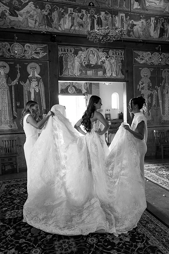 Bride getting ready as bridesmaids bustle her wedding dress, lace train and veil flowing beneath a chandelier in an ornate church interior