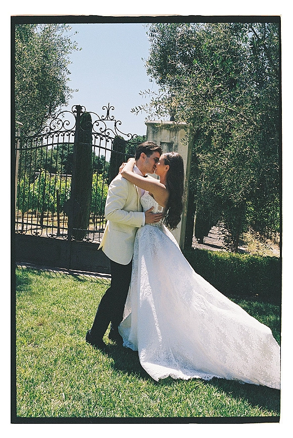 Wedding kiss portrait of bride and groom kissing, her strapless lace gown with long train beside a wrought iron gate on a sunny lawn