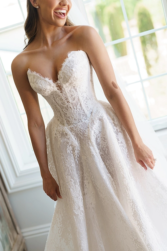 Wedding dress with strapless lace bodice and sweetheart neckline, pearl stud earrings and engagement ring by bright window light