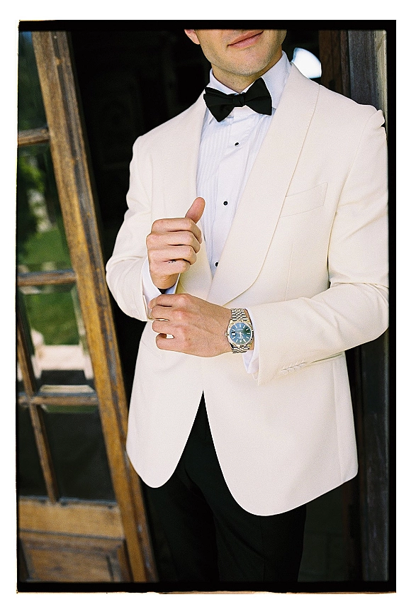 Groom portrait in a white tuxedo jacket with black bow tie, adjusting cuff buttons and wristwatch while standing in a doorway