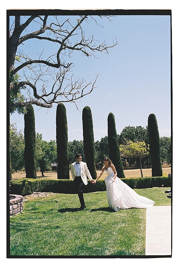 Couple portrait of bride and groom holding hands, her strapless gown with long train beside him in white tux on manicured garden lawn