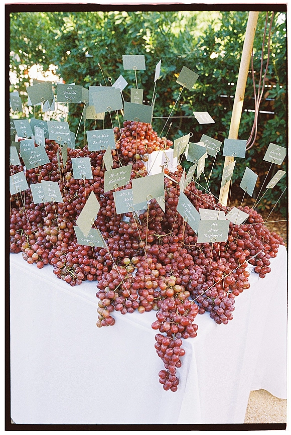 Escort card display with grape escort cards on wire picks, calligraphy place cards on white linen in a wooden frame amid garden foliage