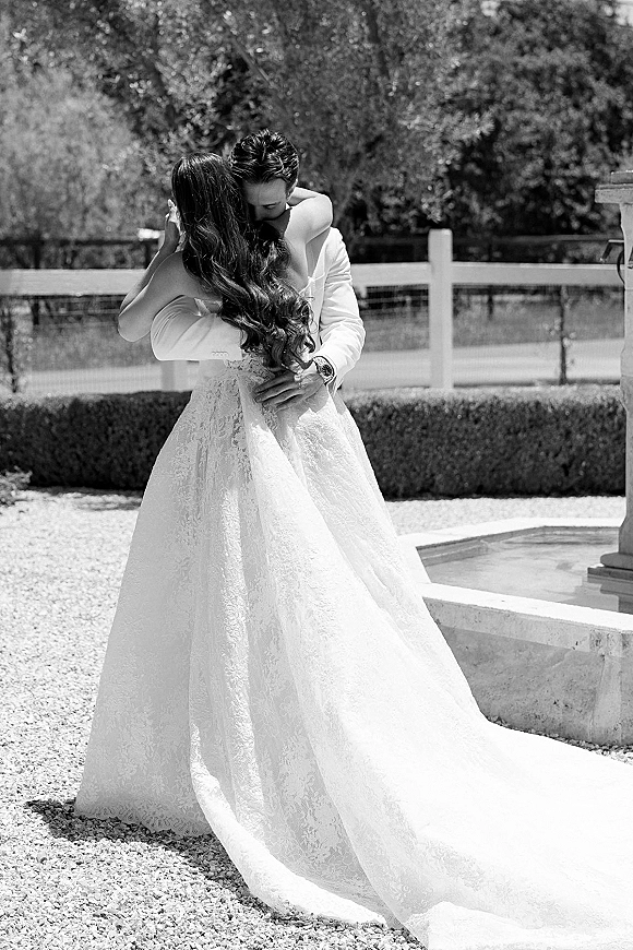 Wedding couple portrait of bride and groom embrace on a gravel garden path, her strapless lace dress with long train trailing behind near a fountain