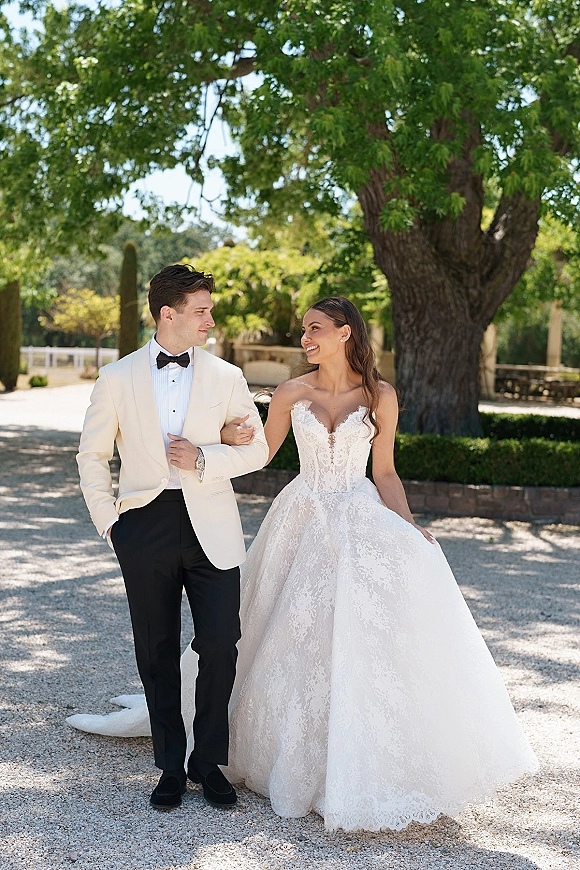 Couple portrait of bride holding groom’s arm as they walk a sunny garden path, her strapless lace dress and his white jacket with bow tie