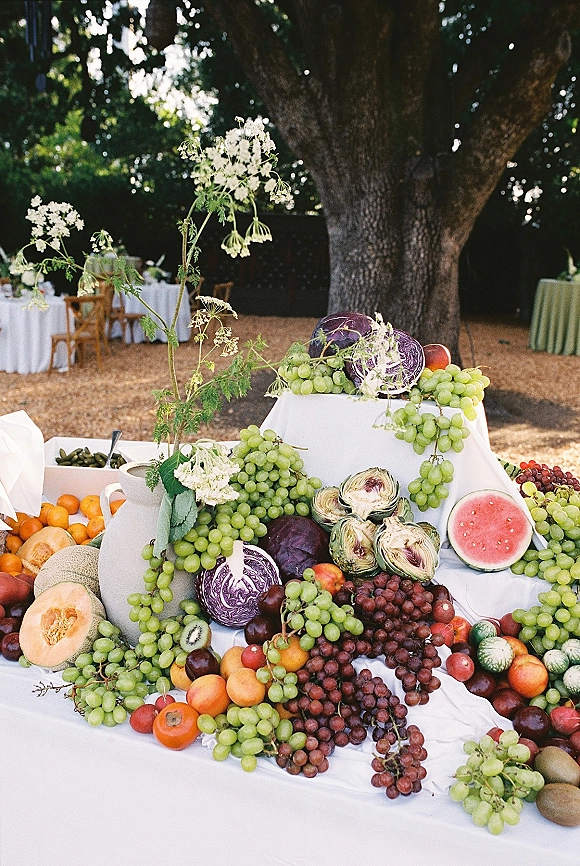 Wedding fruit display with grapes, watermelon, and citrus arranged on white linens, accented with wildflower stems on an outdoor lawn under a large tree