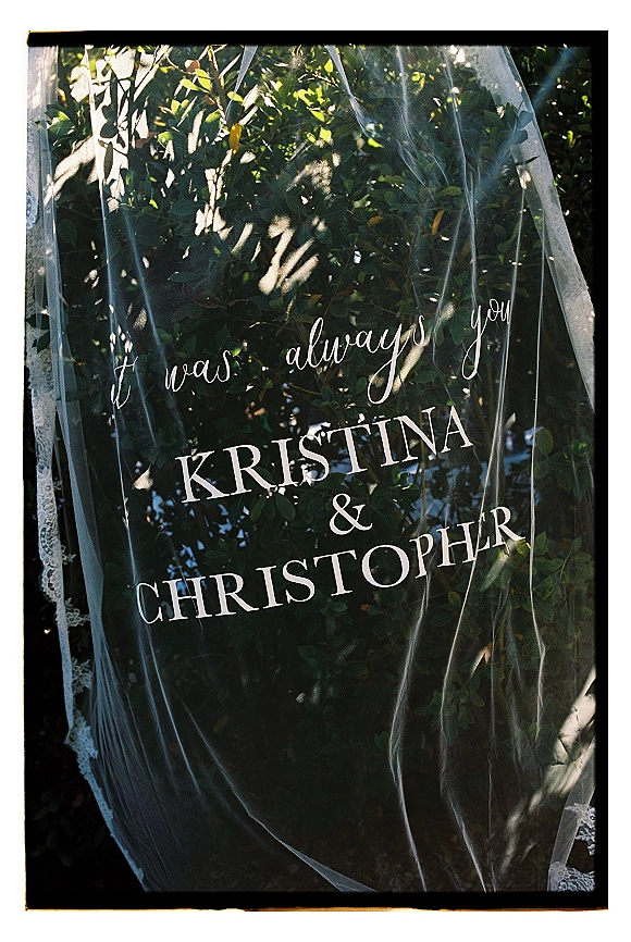 Wedding welcome sign in clear acrylic with calligraphy lettering, lace veil draped over it against sunlit green foliage and shadows