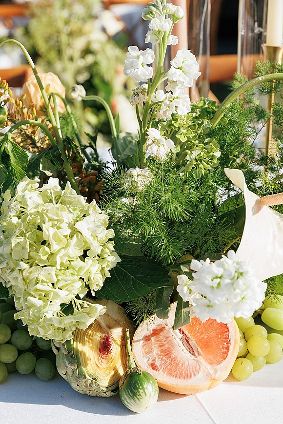 Wedding centerpiece featuring a green and white centerpiece with hydrangea, white flowers, greenery, fruit and candles on a sunlit table