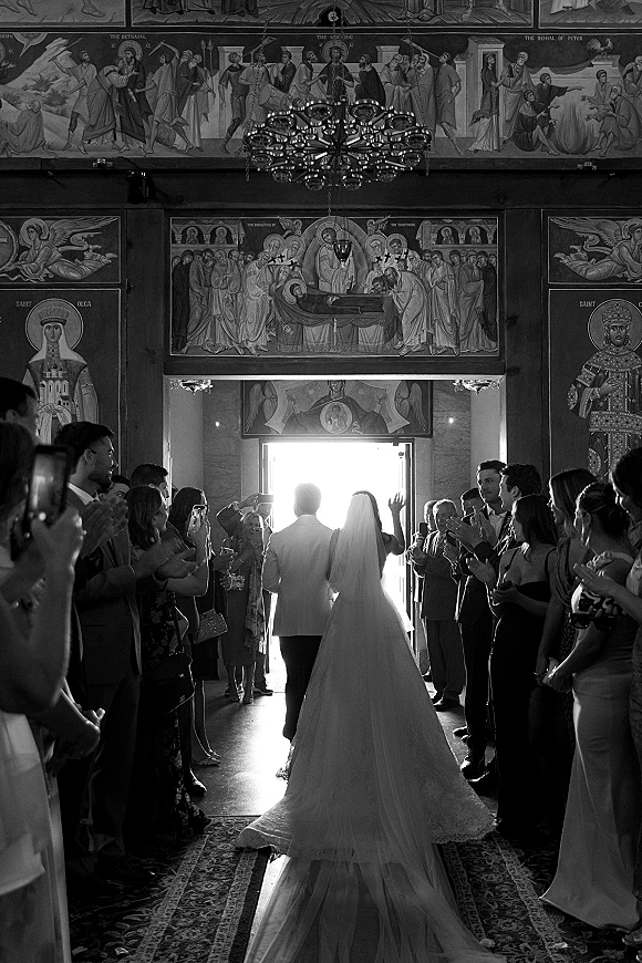 Wedding recessional as bride and groom walking out down a church aisle, her long veil and train backlit by the doorway as guests clap
