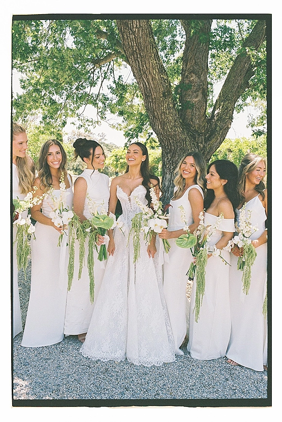 Bridesmaids portrait of the bride with bridesmaids in white dresses holding cascading orchid bouquets on a sunlit gravel path by a large tree