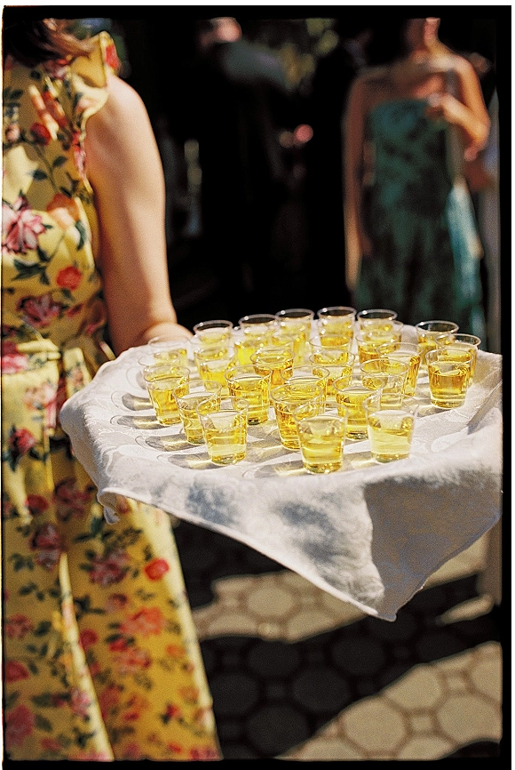 Wedding drink service with a tray of shot glasses filled with yellow drinks on white linen, held by a guest in a floral dress at reception