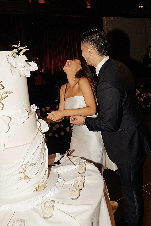 Wedding cake cutting as the bride and groom cutting cake, laughing beside a tall white cake with sugar flowers and votive candles.
