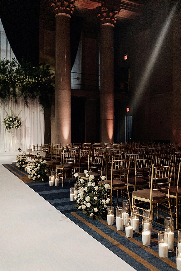 Ceremony aisle decor with a white aisle runner, gold chiavari chairs, and candle-lined glass cylinders in a columned ballroom setting