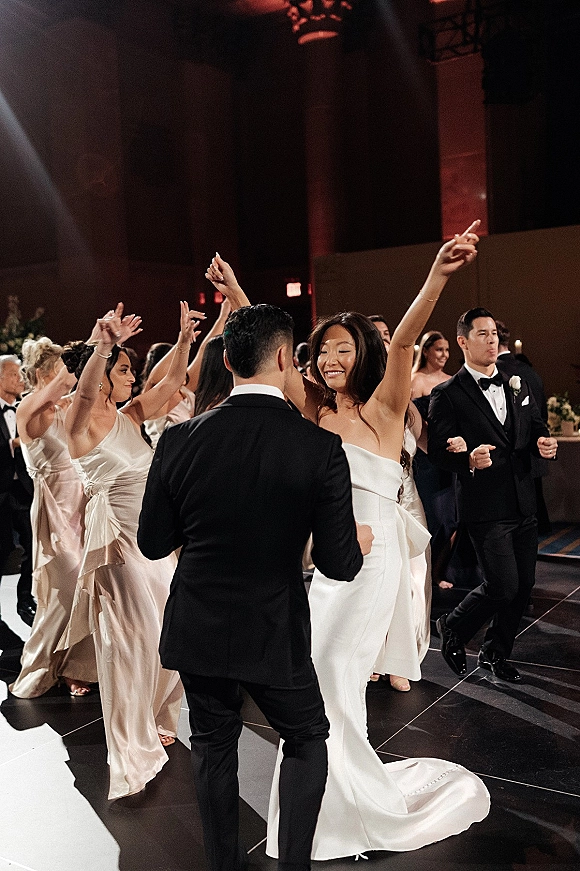 Wedding dance floor packed with bride in a strapless gown and groom in tuxedo dancing with bridesmaids in satin dresses under candlelit ballroom lights