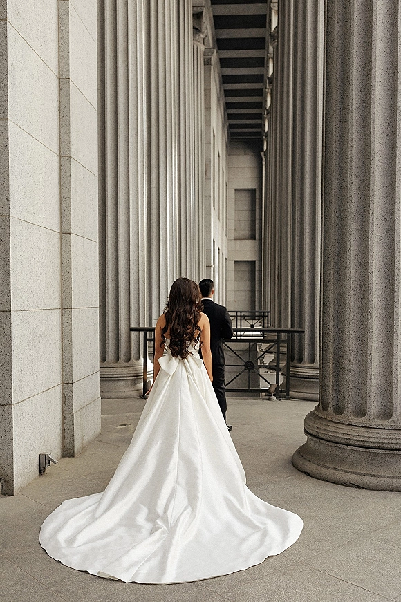 Wedding first look as bride approaches groom from behind, satin gown with long train and bow detail in stone colonnade walkway