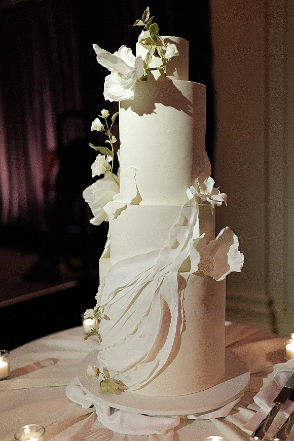 Wedding cake with white fondant and sugar flowers, greenery, and votive candles on a stand against a dark wood reception wall