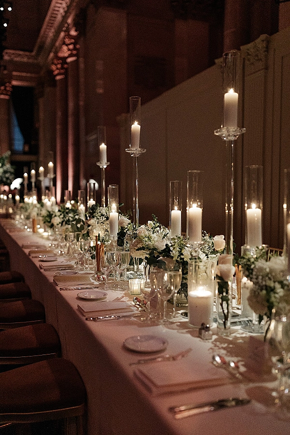 Reception tablescape with long head table decor featuring taper and pillar candles, low white florals, and greenery runner in a dim arched hall