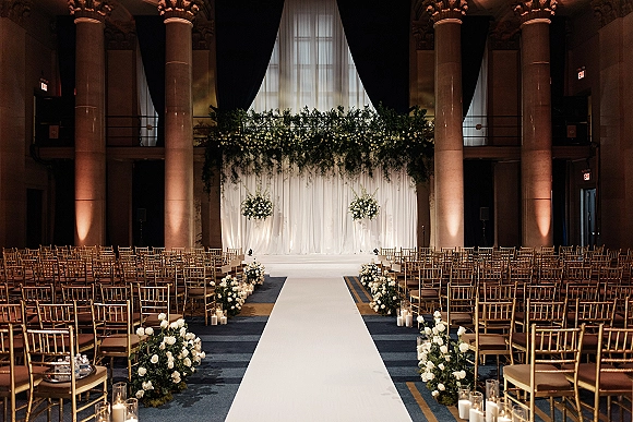 Ceremony setup for an indoor wedding ceremony with white aisle runner, gold Chiavari chairs, and candlelit florals in a grand ballroom