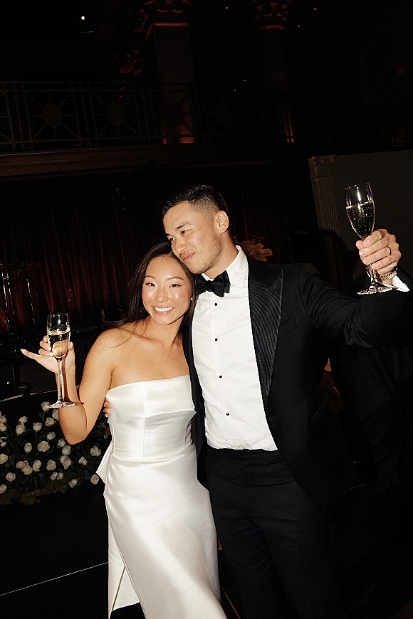 Couple portrait of bride in a strapless satin wedding dress and groom in black tuxedo raising champagne flutes in a dim reception room