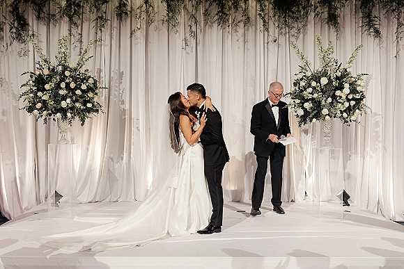 Wedding kiss as bride in strapless dress and long veil kisses groom in black tux on a white-draped indoor stage with florals