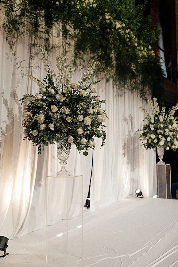 Wedding ceremony decor with white draped ceremony backdrop, white rose arrangements with greenery in crystal urn vases on clear acrylic pedestals.