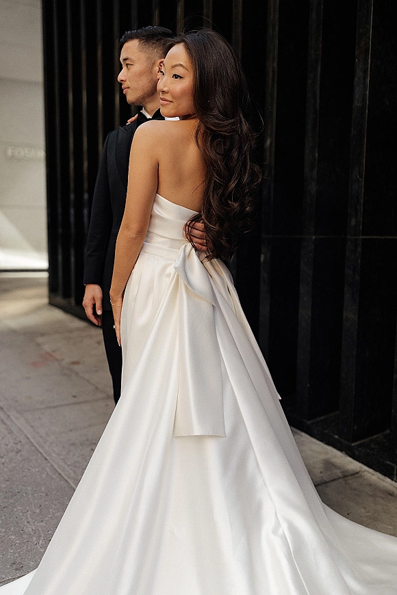 Couple portrait of bride and groom embracing, her strapless satin wedding dress with oversized bow against dark urban wall panels