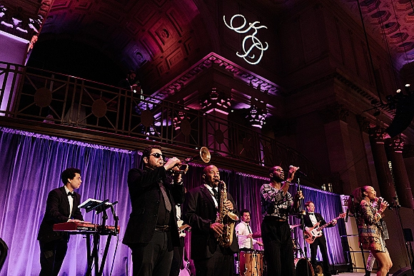 Wedding reception band performing as a live wedding band with horn section and microphones on stage beneath ballroom balcony and columns